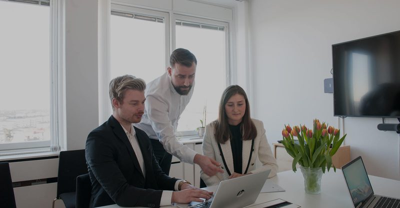 Tre medarbetare vid ett mötesbord med utsikt över Stockholm, en stående man i vit skjorta lutar sig fram och pekar mot laptopskärmen framför sina två sittande kollegor