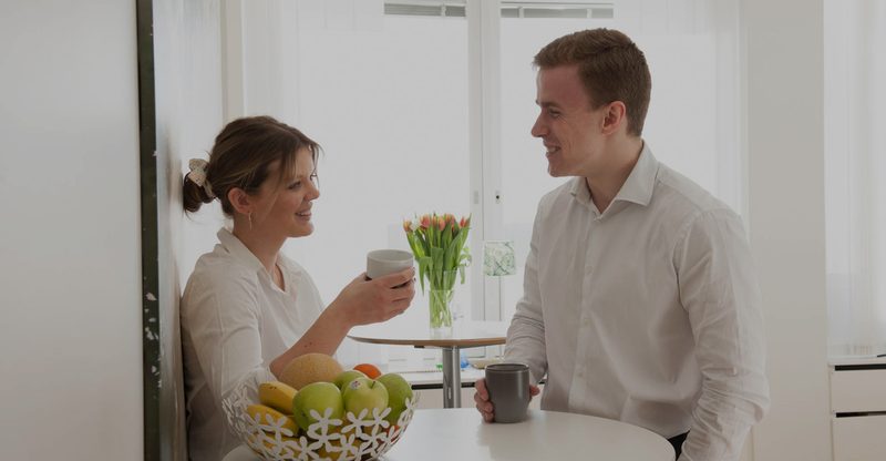 Två unga medarbetare i vita skjortor fikar och skrattar vid ett runt bord i kontorsköket, med kaffemuggar, fruktkorg och tulpaner på bordet