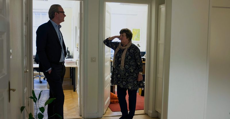 Två medarbetare står och samtalar i dörröppningarna mellan två kontorsrum, en gråhårig man med glasögon i mörk kavaj och en kvinna i mönstrad blus som lutar armen mot dörrkarmen